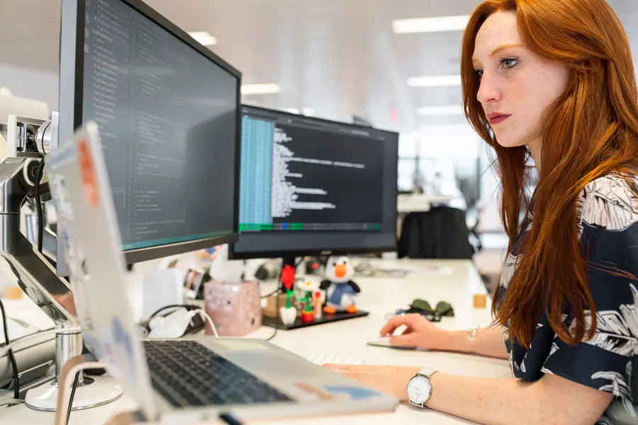 Femme développeuse examinant son code devant plusieurs écrans, représentant les compétences en webdesign et l'optimisation de l'expérience utilisateur pour une meilleure performance en ligne.