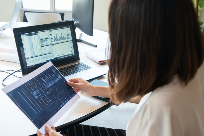 Une femme devant un ordinateur affichant des statistiques, tenant en main une impression des données, illustrant l'analyse des campagnes de netlinking pour un SEO impactant grâce à un portefeuille de backlinks équilibré.