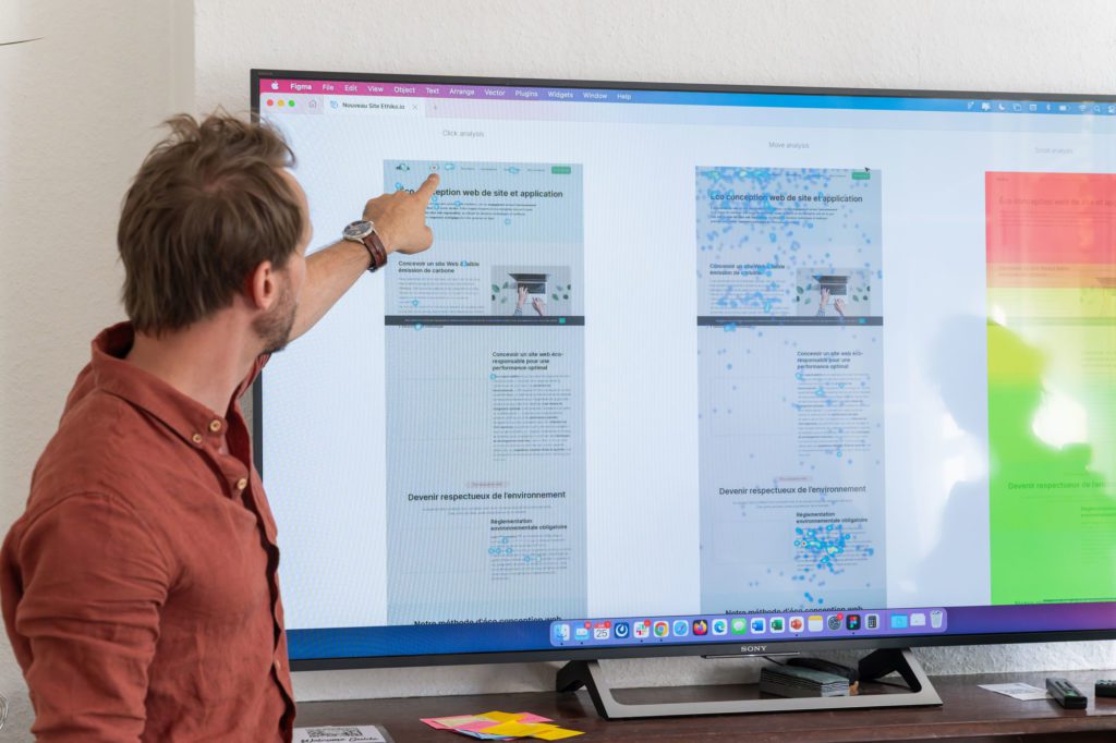 Un homme en chemise rouge pointe du doigt un grand écran d'ordinateur posé sur une cheminée en bois. L'écran affiche une interface Figma avec des pages web intitulées "Éco conception web de site et application" et "Éco conception web de site et application". Les pages montrent des informations sur l'optimisation des performances et la réduction des émissions de carbone, avec des graphiques et du texte.