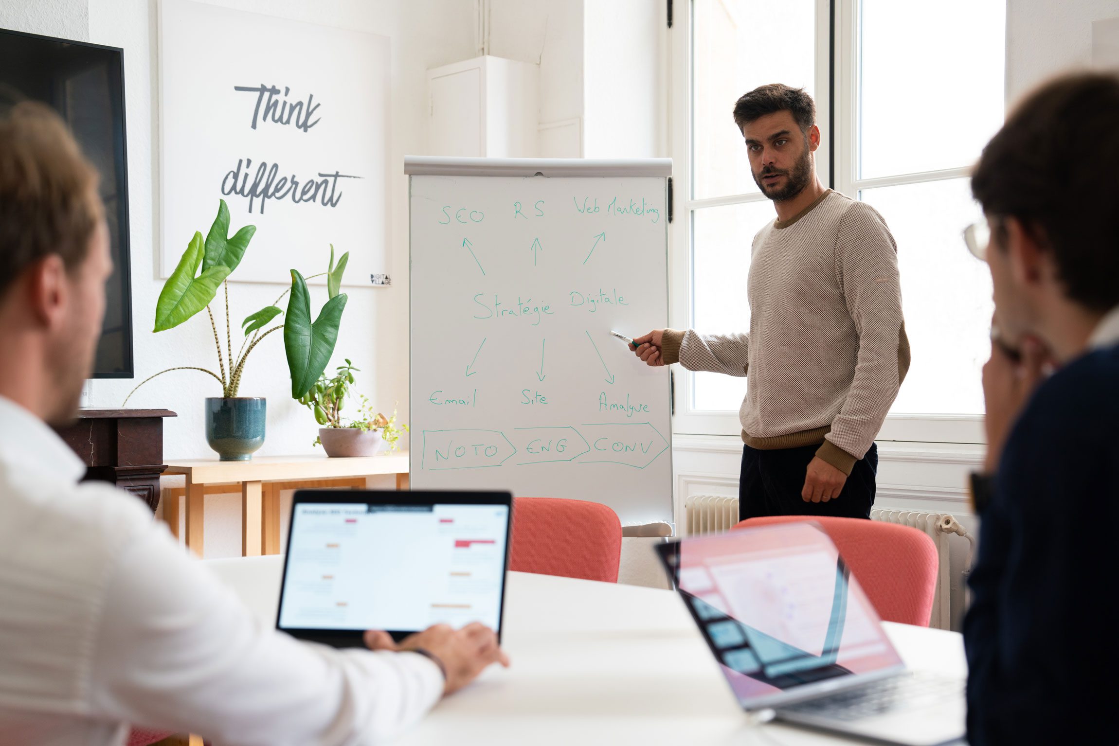 Un homme en pull beige présente un tableau blanc dans une salle lumineuse, expliquant une stratégie digitale avec des termes comme "SEO", "RS", "Web Marketing", "Email", "Site", "Analyse", "Noto", "Eng", et "Conv". Deux personnes assises à une table avec des ordinateurs portables l'écoutent. Une plante verte est posée sur une table à gauche, et une inscription "Think different" est visible sur le mur.